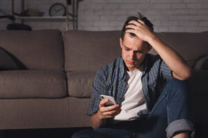 a teen boy stares at his phone while holding his head nervously, one of many signs of potential mental health issues in teens
