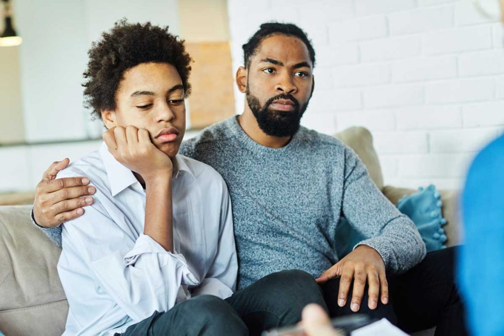 a son looks annoyed but a father looks attentive as he listens to a therapist talk about how he can help his son with a mental illness