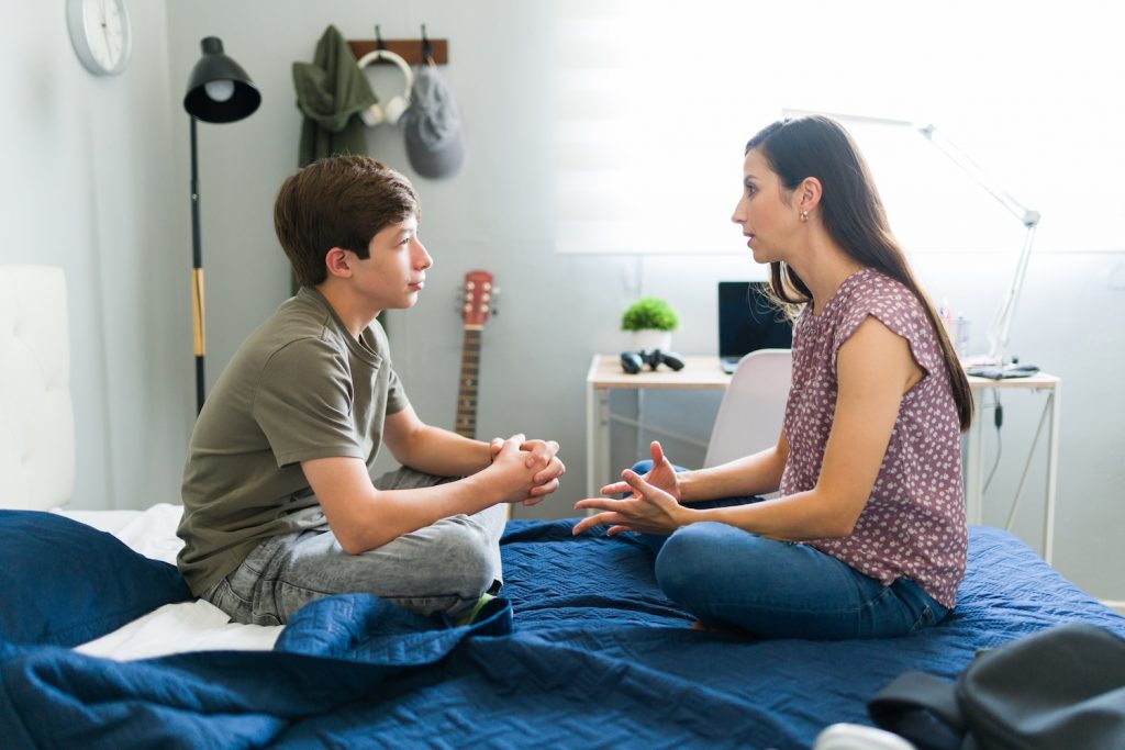 Mother talking with her teenage son while sitting on his bed.