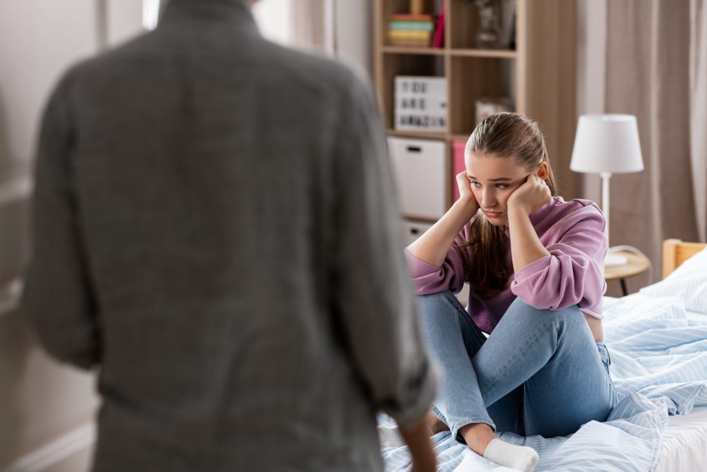 Upset teenage girl sitting on her bed while an adult speaks to her.