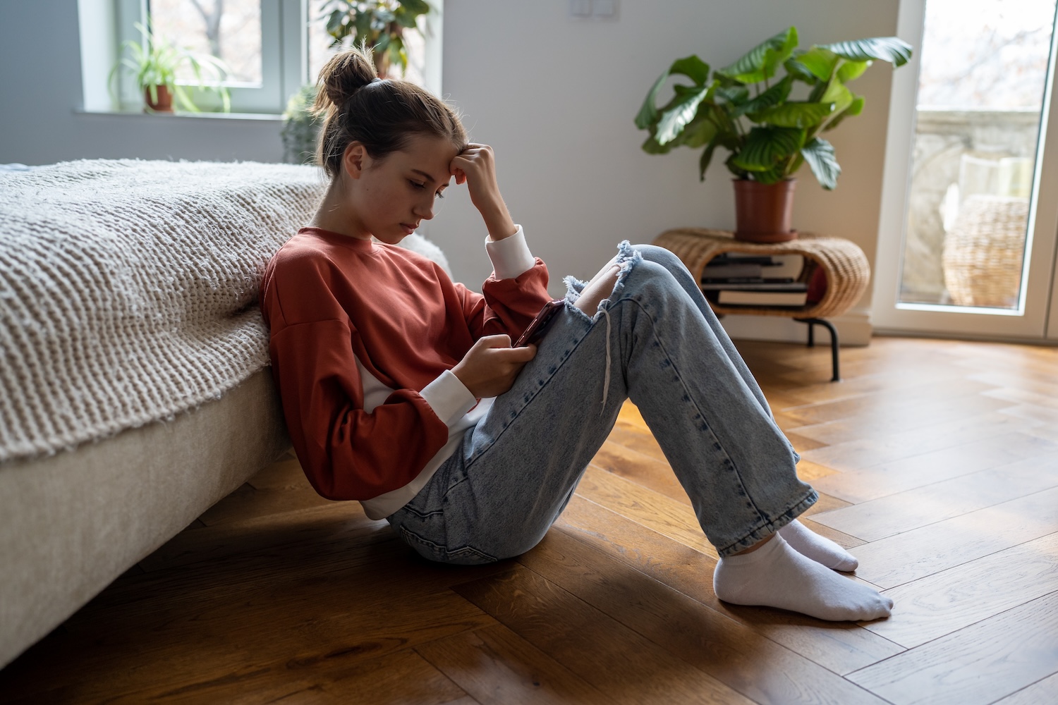 A young person sits on the floor beside a bed, leaning forward while looking at a phone. They rest one hand on their forehead and appear deep in thought. Natural light comes through nearby windows, and houseplants and books are visible in the background.