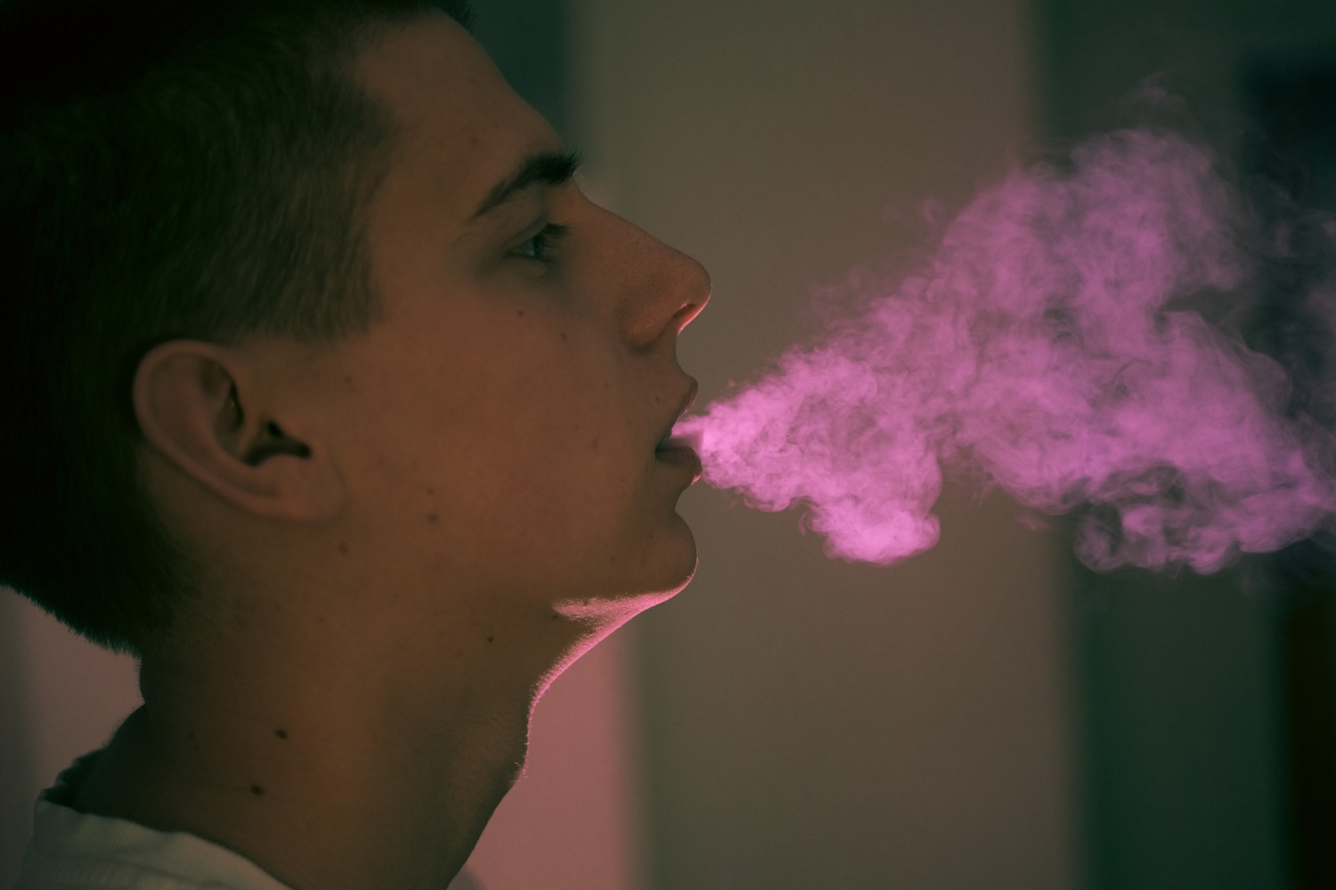 A side profile of a person exhaling a thick cloud of pink vapor in a dimly lit room.