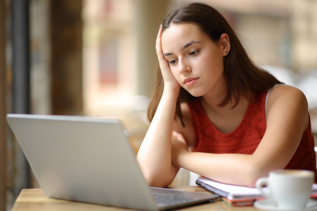 A young woman sits at a table with her head resting on her hand, looking tired or discouraged as she stares at a laptop screen. A notebook and a cup of coffee are beside her.