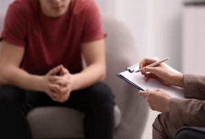 A person sits leaning forward with their hands clasped while a therapist writes notes on a clipboard during a counseling session.